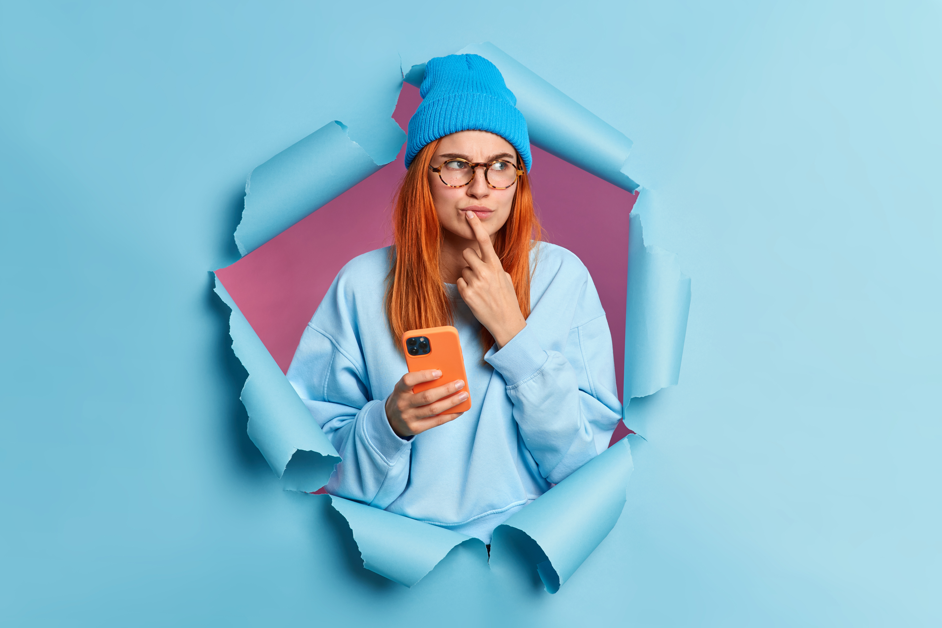 pensive-redhead-woman-holds-mobile-phone-with-thoughtful-expression-keeps-index-finger-near-lips-browses-internet-thinks-received-message-wears-hat-jumper-breaks-through-blue-paper-wall.jpg